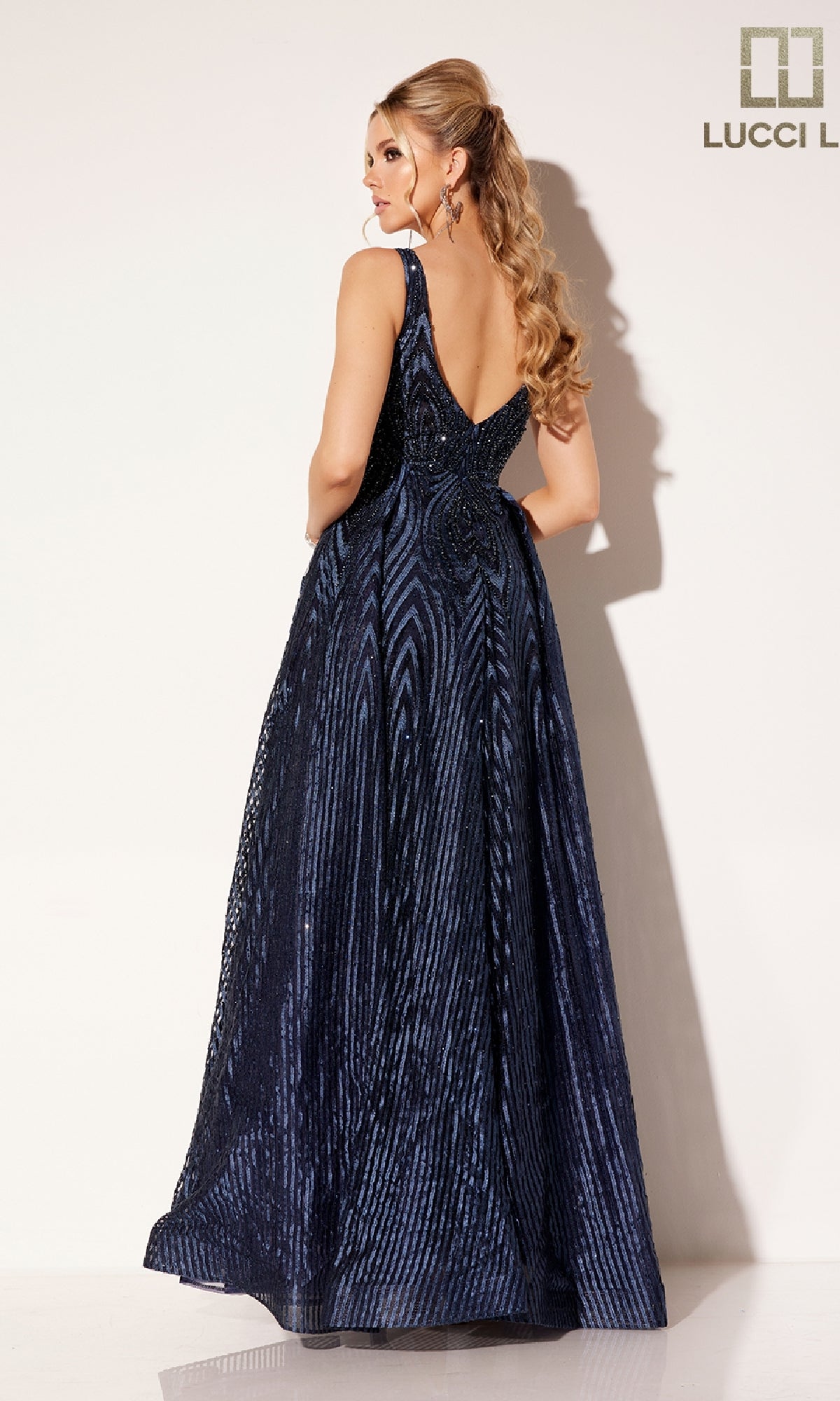 A woman with long wavy hair models the Lucci Lu C8074, a floor-length, sleeveless navy blue prom dress with a deep V-back, standing against a plain light background.